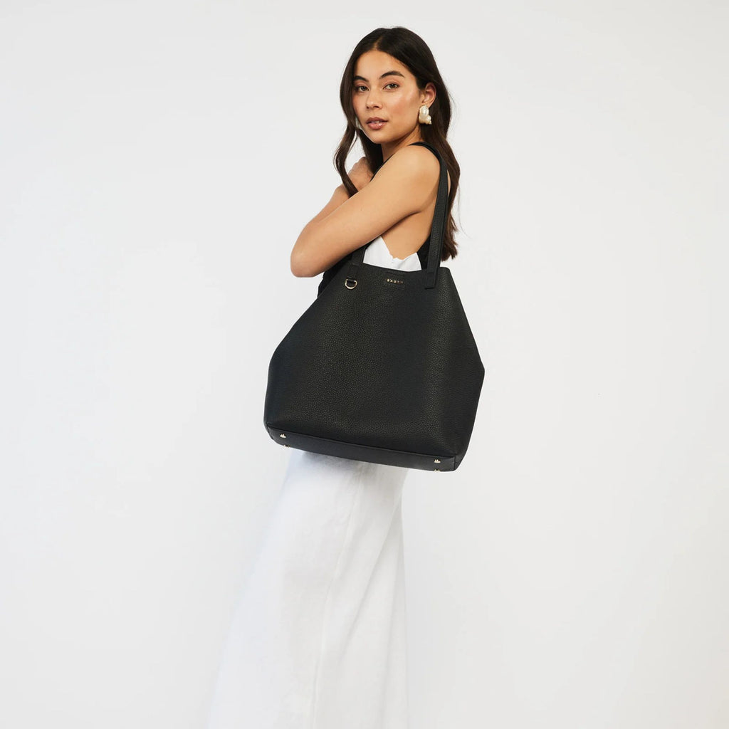 A woman wearing all white with a Black leather tote bag with long shoulder strap on her shoulder on a white background