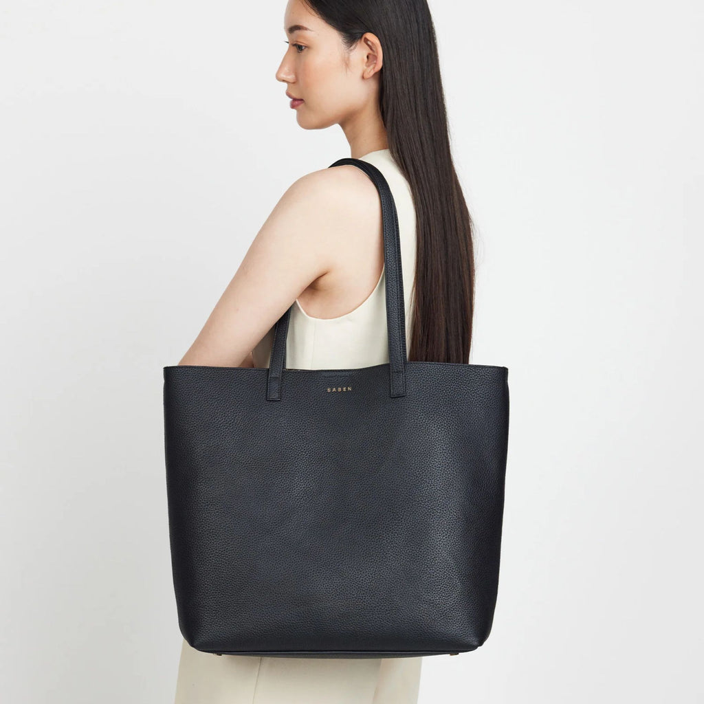 A woman wearing all white with a Black leather tote bag with long shoulder strap on her shoulder on a white background