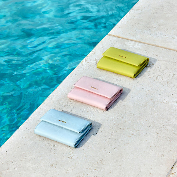 Three folded wallets in pastel blue, pink, and yellow-green are arranged diagonally on a light stone surface next to a swimming pool.