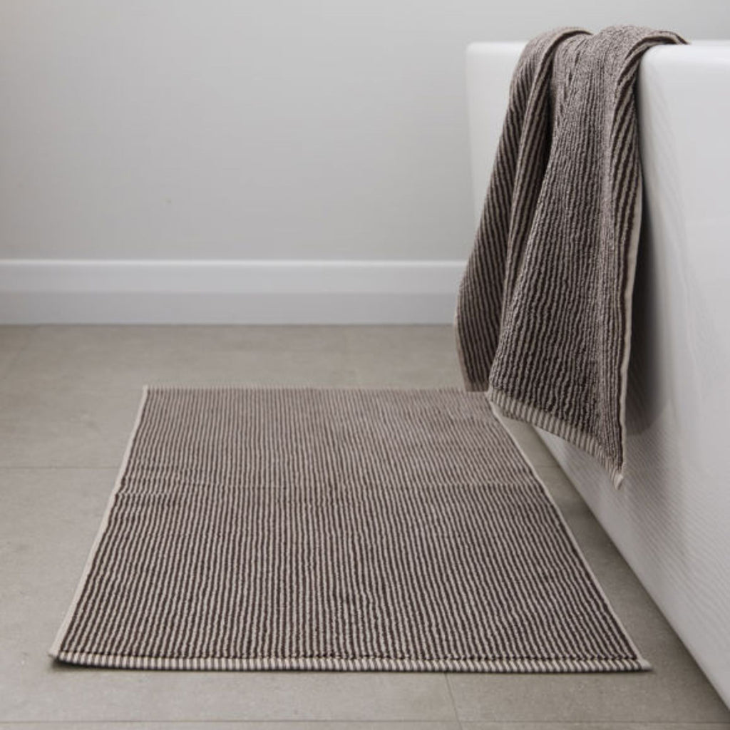 Brown textured bath mat on a light gray floor next to a white bathtub with a brown towel draped over the edge.