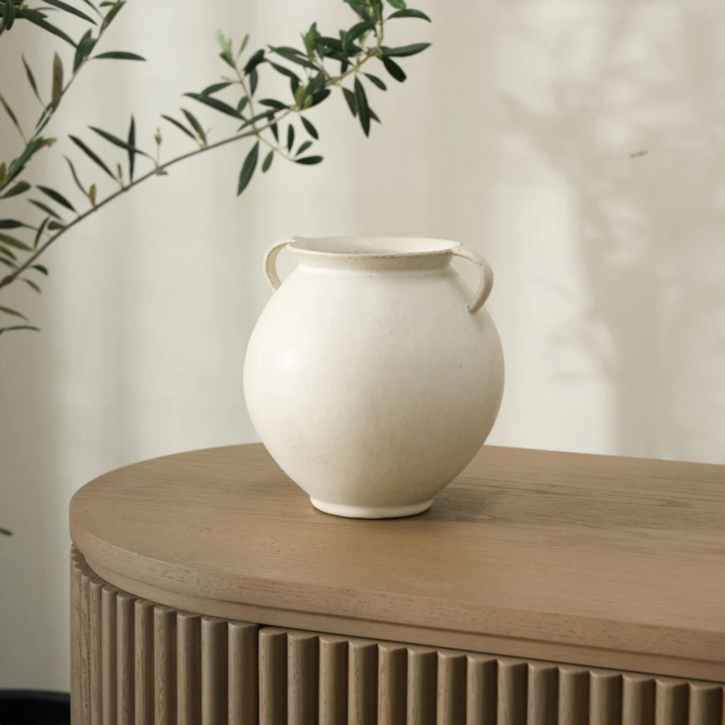 White ceramic vase with handles on a wooden surface with a blurred plant in the background