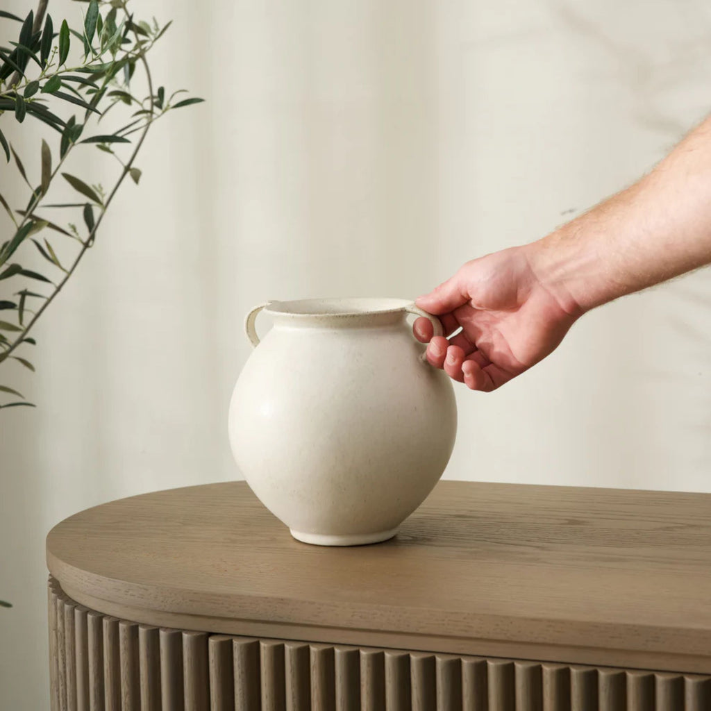 Person holding a white ceramic vase on a wooden surface with a plant in the background