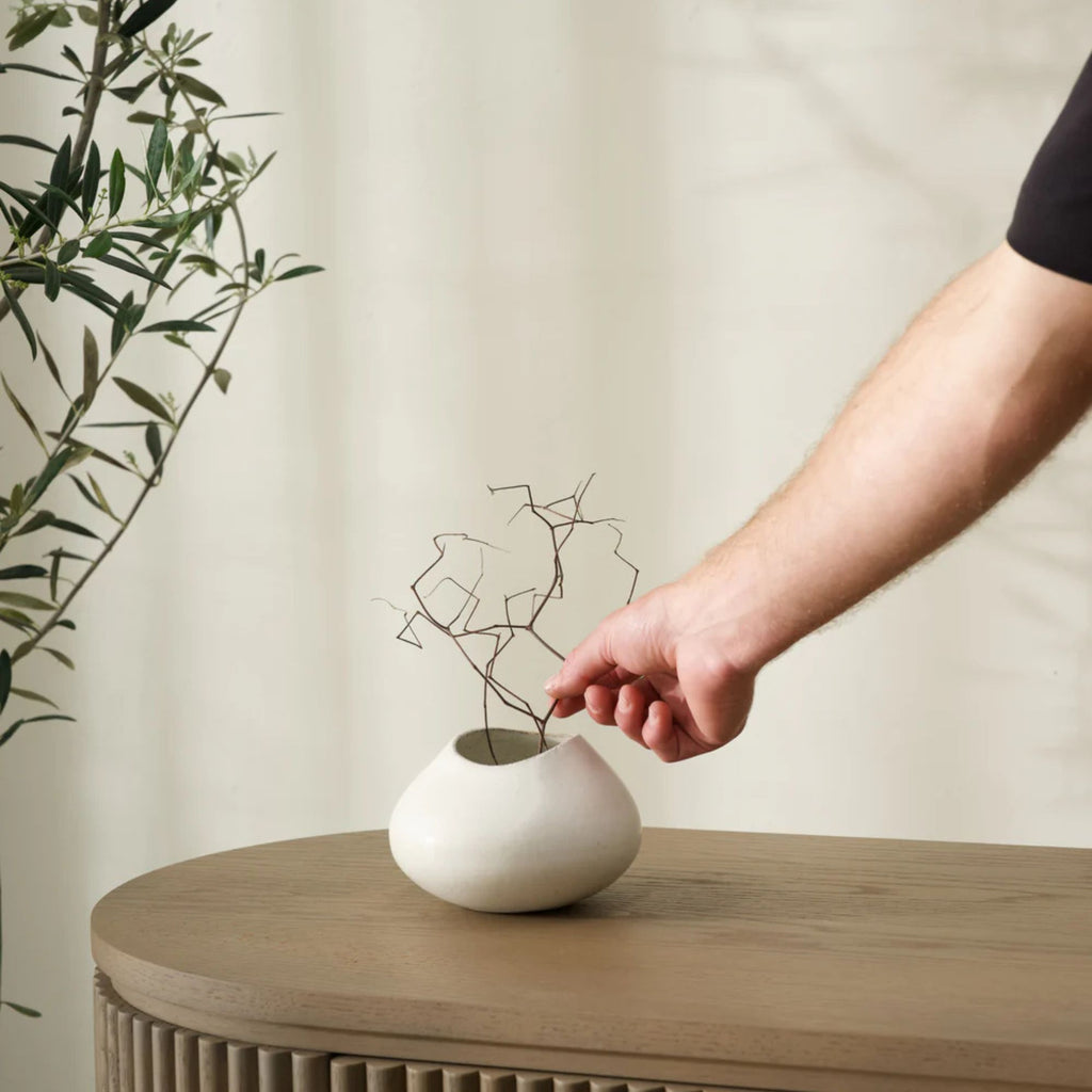 Person placing a branch into a small white vase on a wooden surface with a neutral background