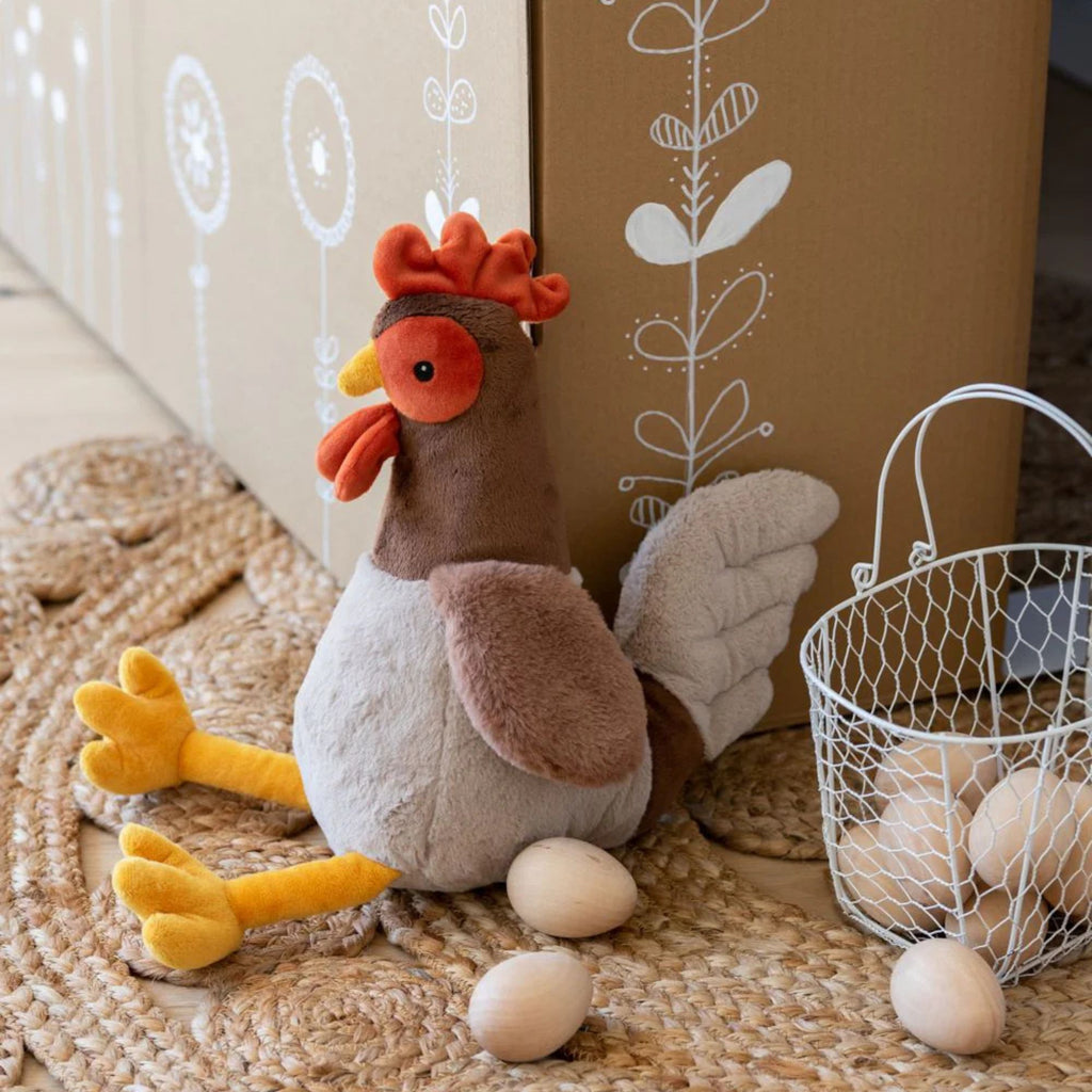 a soft toy rooster sitting next to a basket of eggs on a natural rug