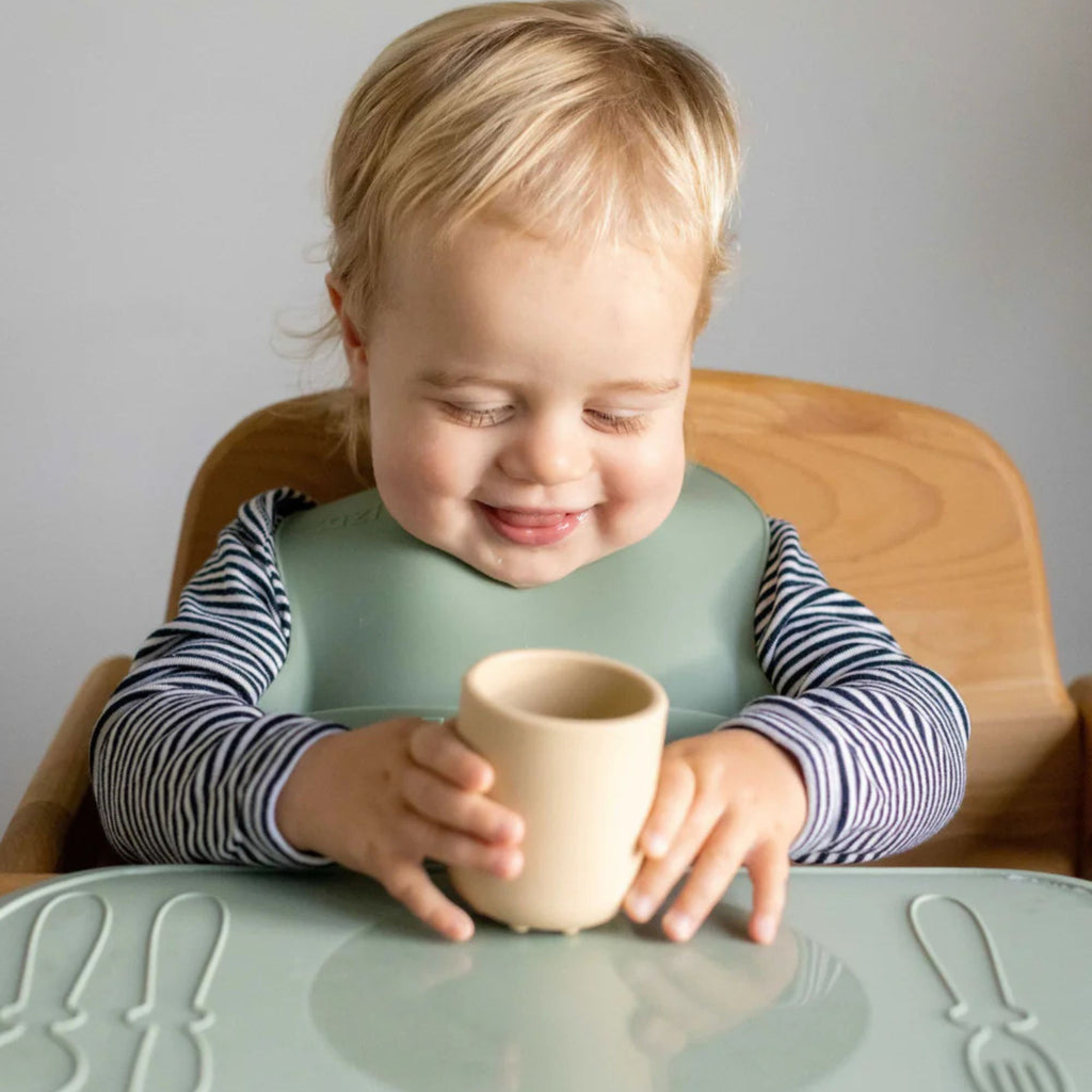 Child sitting at a table with a cup, wearing a bib, on a neutral background