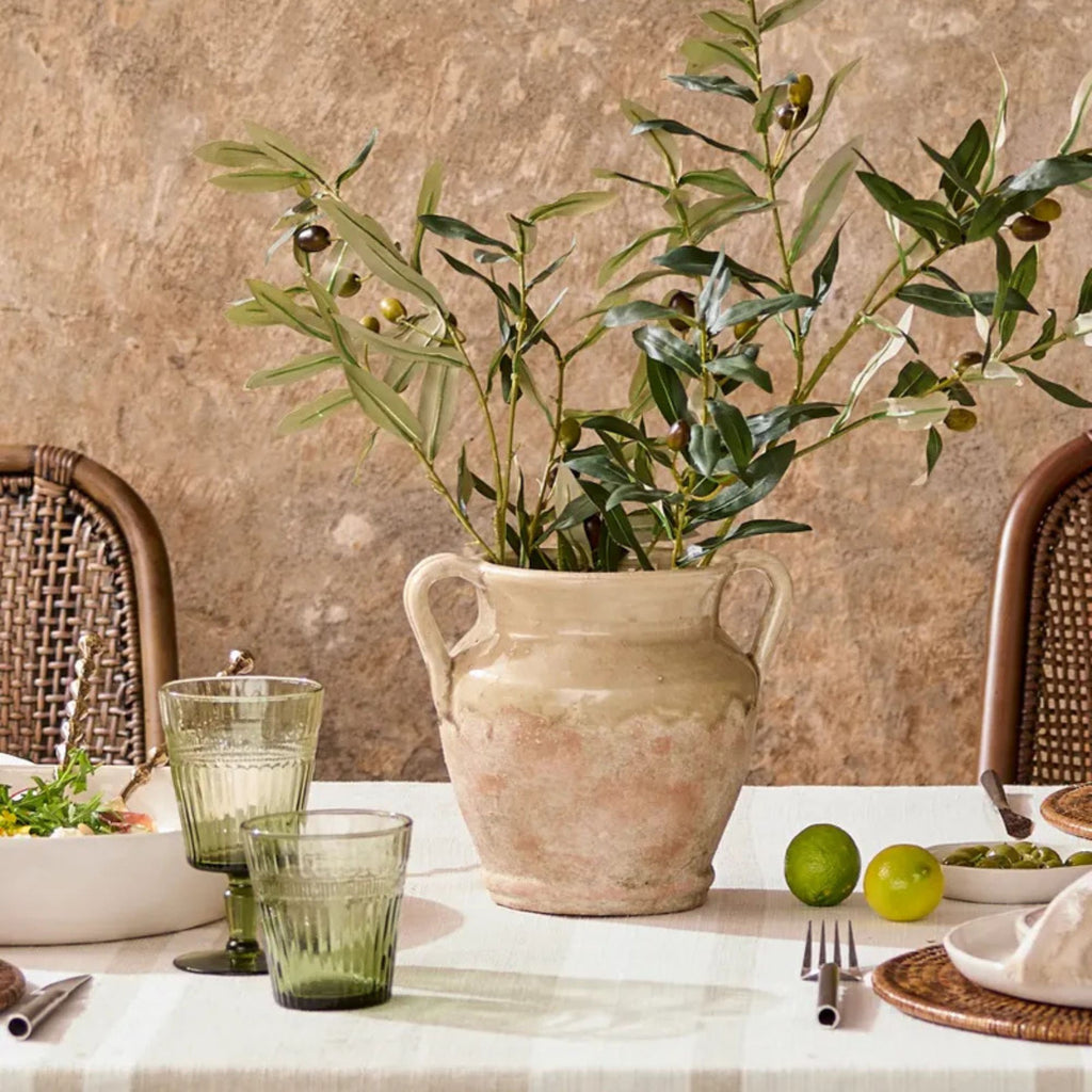 Table setting with a terracotta vase, green glasses, and limes against a textured wall.