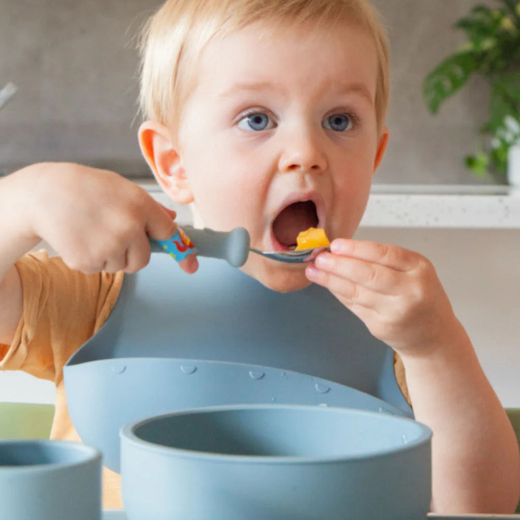 Child eating with a spoon and wearing a blue bib indoors.