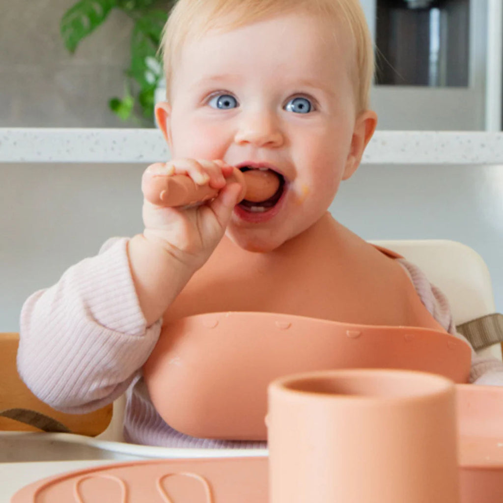 Baby sitting in a high chair with a pink tray, eating with a spoon.