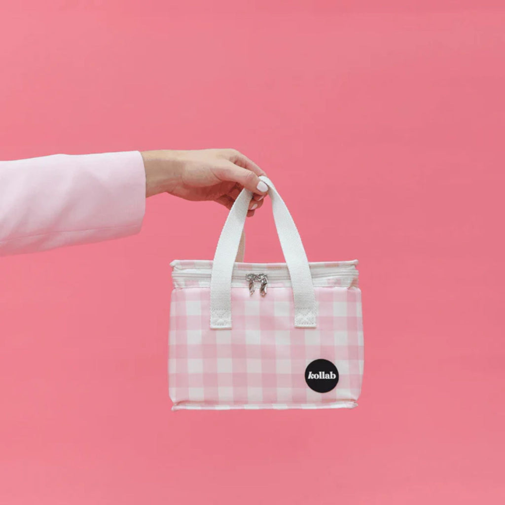 A pink checked lunch bag with white straps and Kollab branding held by an arm against a pink wall