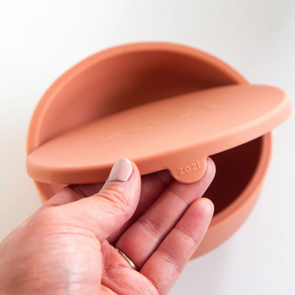 Terracotta-colored silicone bowl with 'zazi' branding held by a hand on a white background