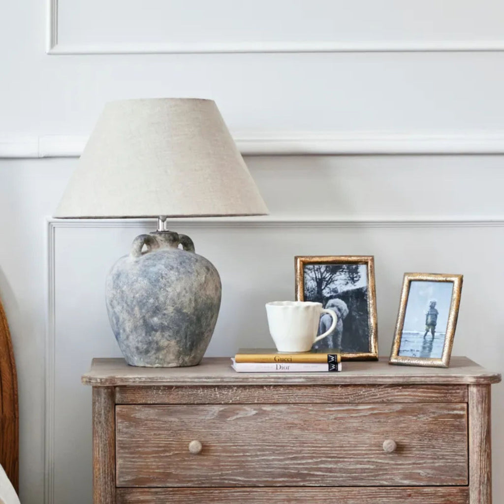 Wooden dresser with a lamp, vase, mug, and books against a white wall.