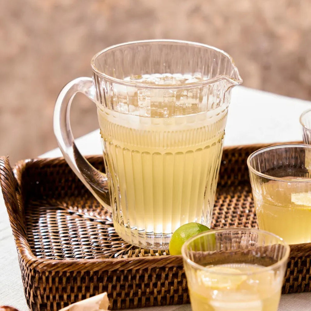 Clear pitcher and glasses filled with a light yellow beverage on a woven tray.