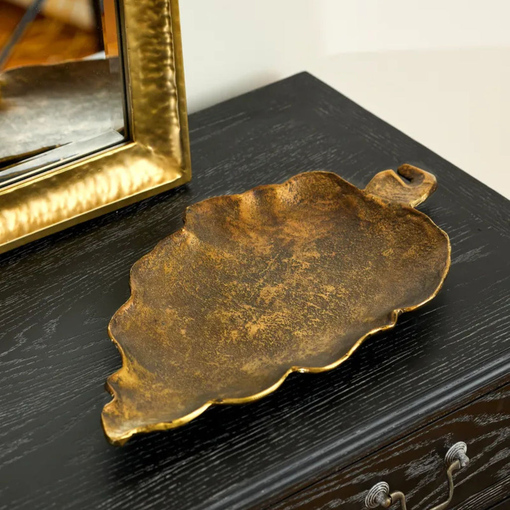 Bronze leaf-shaped tray on a dark wooden surface with a gold-framed mirror in the background.