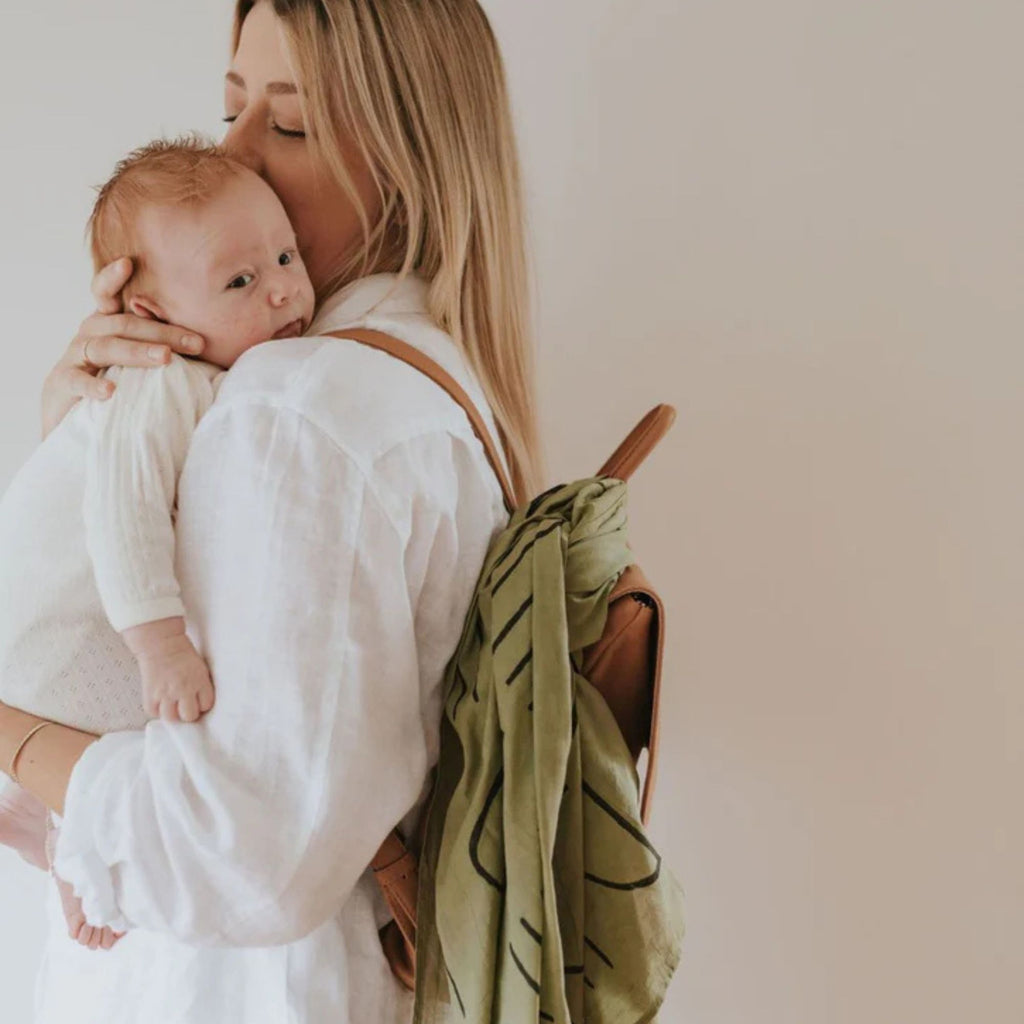 Woman holding a baby and carrying a green backpack with a leaf pattern on a plain background