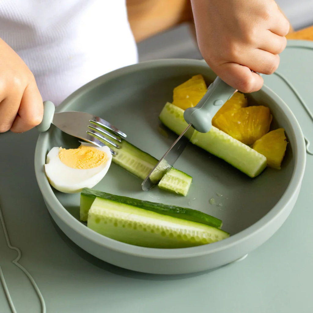 Children using cutlery to eat sliced cucumbers and eggs on a plate.