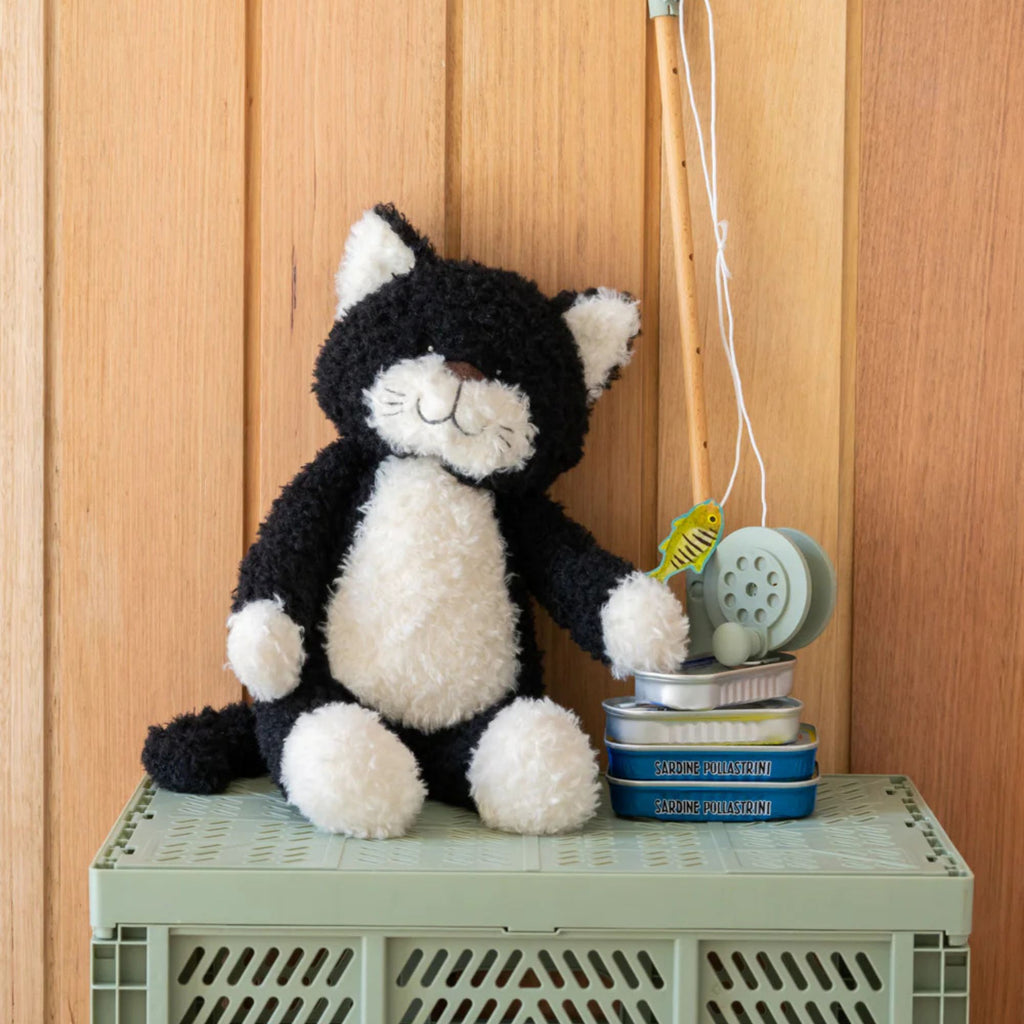 A stuffed toy black and white cat sitting against a wooden background with a fishing rod and sardine tins