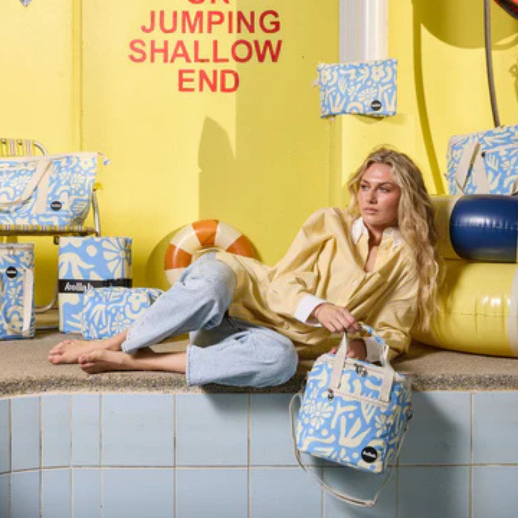 Woman sitting by a pool with 'Jumping Shallow End' sign and blue and white bags.