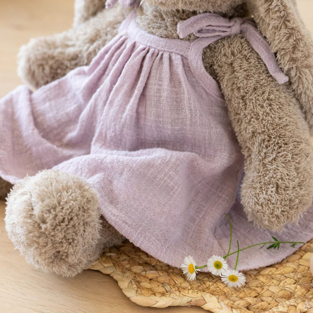 A beige stuffed toy rabbit in a purple linen dress sitting against aa neutral background