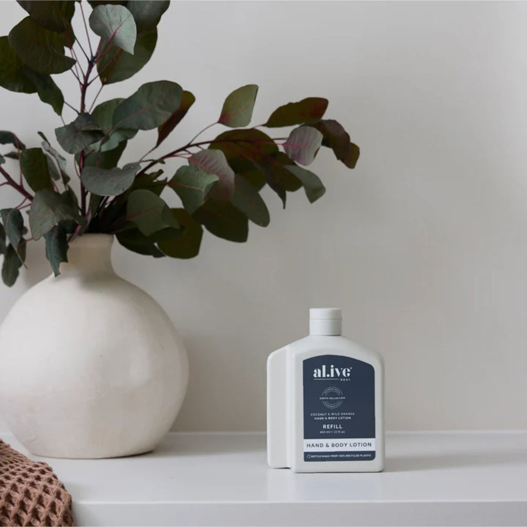 White bottle of al.ive hand and body lotion on a surface with a vase and eucalyptus leaves in the background.