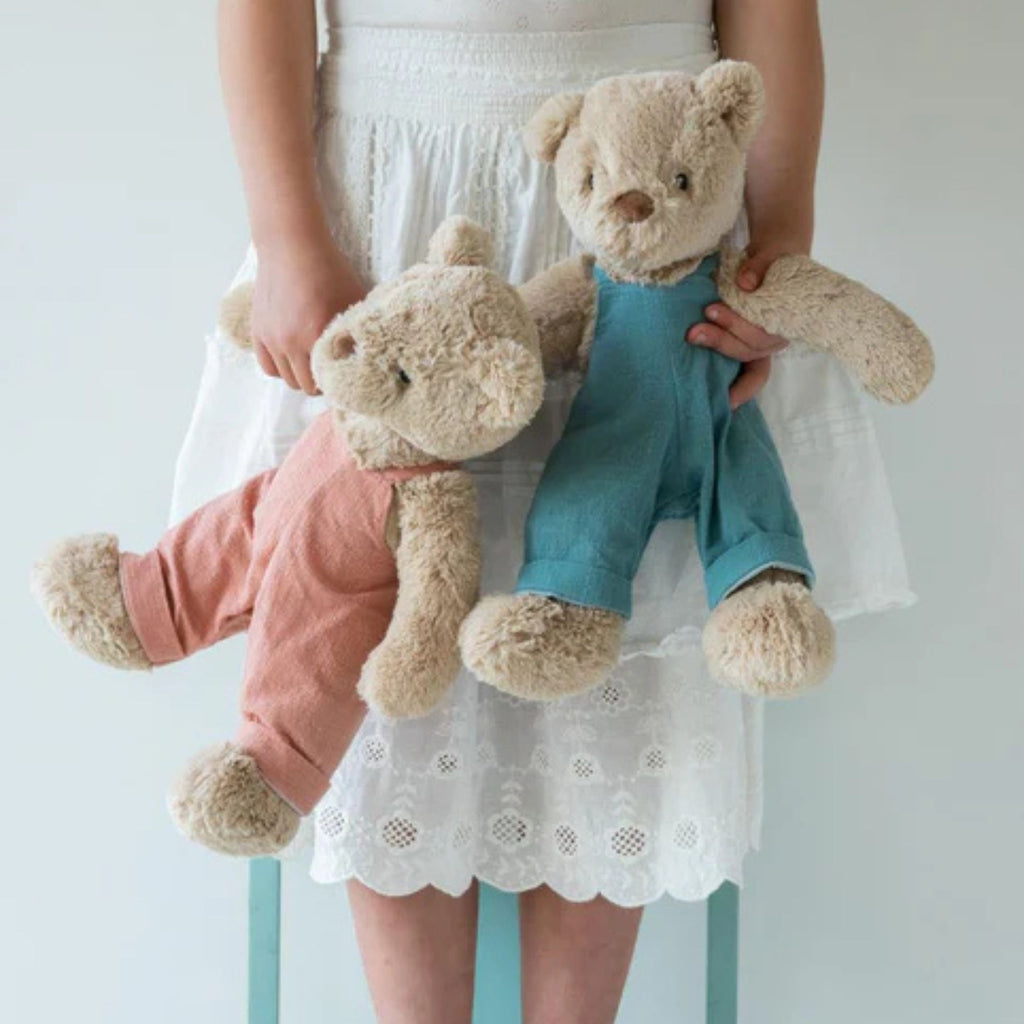 A beige teddy bear in pink linen overalls  and one in blue overalls behing held in each hand by a person