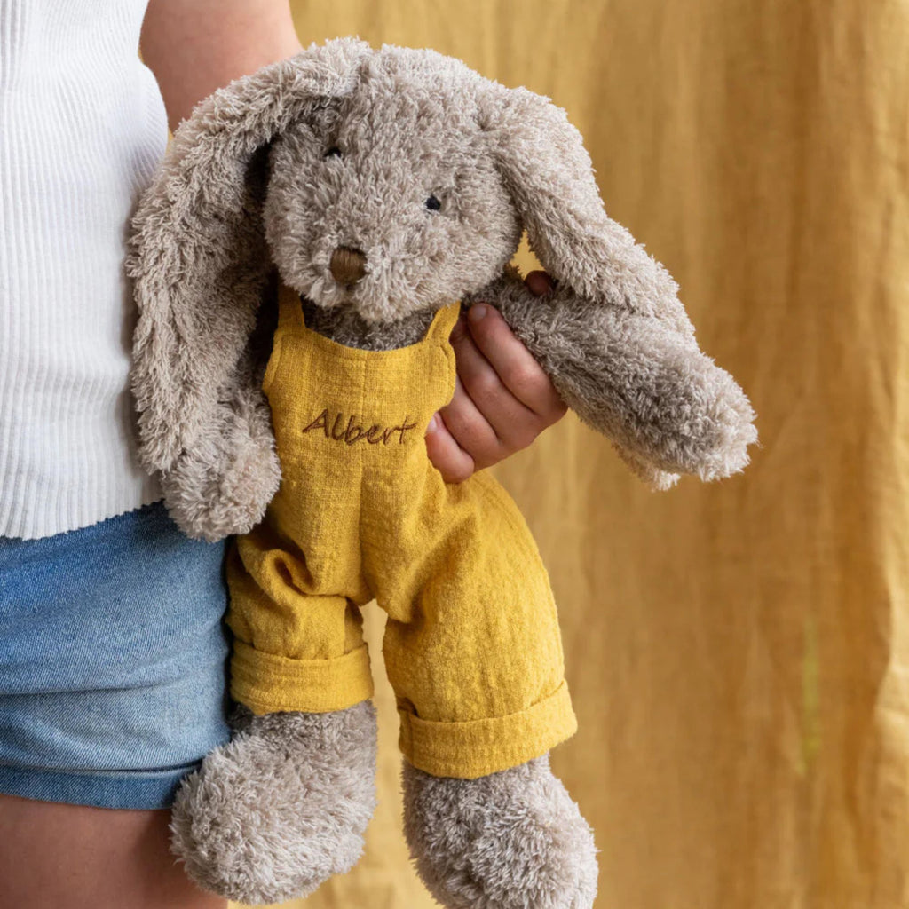 a plush soft toy bunny being held in a persons hand wearing mustard coloured overalls with the name Albert on the front