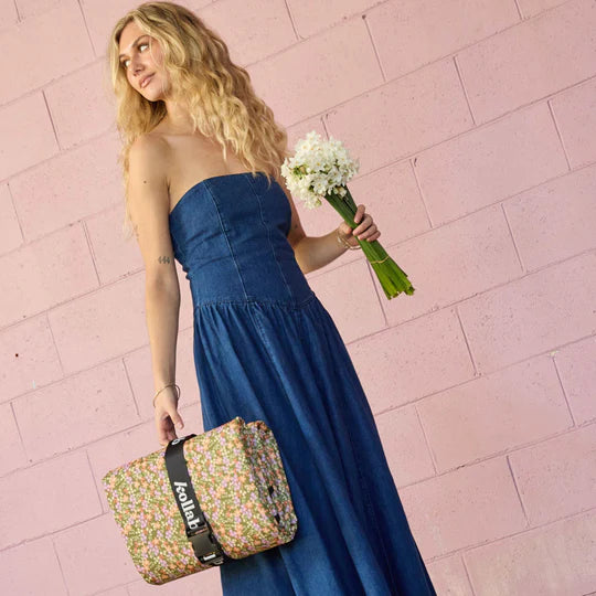 Woman in a blue dress holding a floral bag and bouquet against a pink tiled wall.
