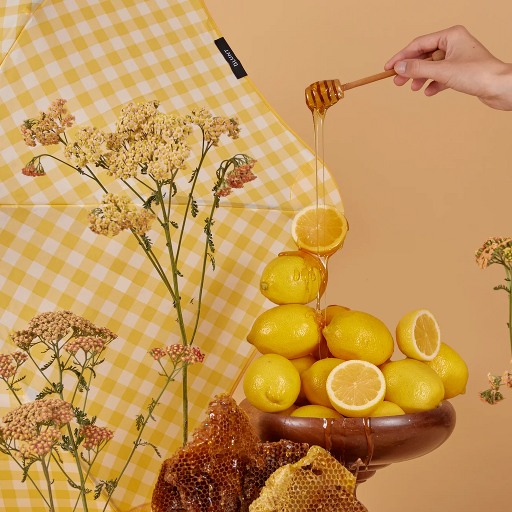 A yellow and white checkered pattern blunt brand umbrella behind home flowers, lemons and a drizzling honey on honey comb against a yellow background 