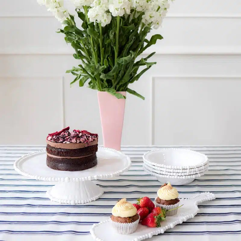 Small chocolate cake, cupcakes with strawberries and cream, and a vase of white flowers on a striped tablecloth.