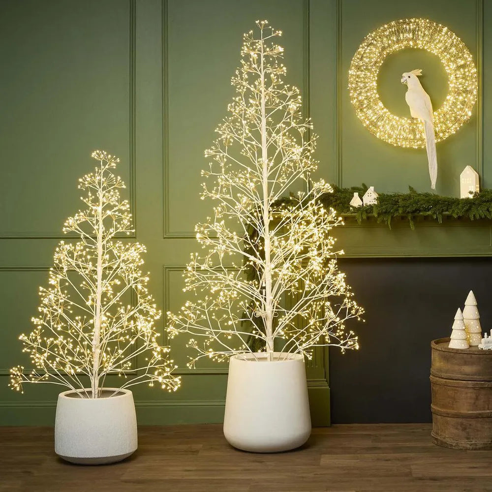 Two potted LED trees with a decorative wreath on a wall in the background.