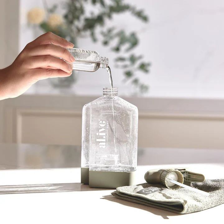 Person pouring liquid from a small container into a larger glass bottle on a light surface with a blurred background.
