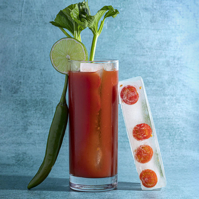 Glass of Bloody Mary with lime, pepper, and ice cube tray on a blue background
