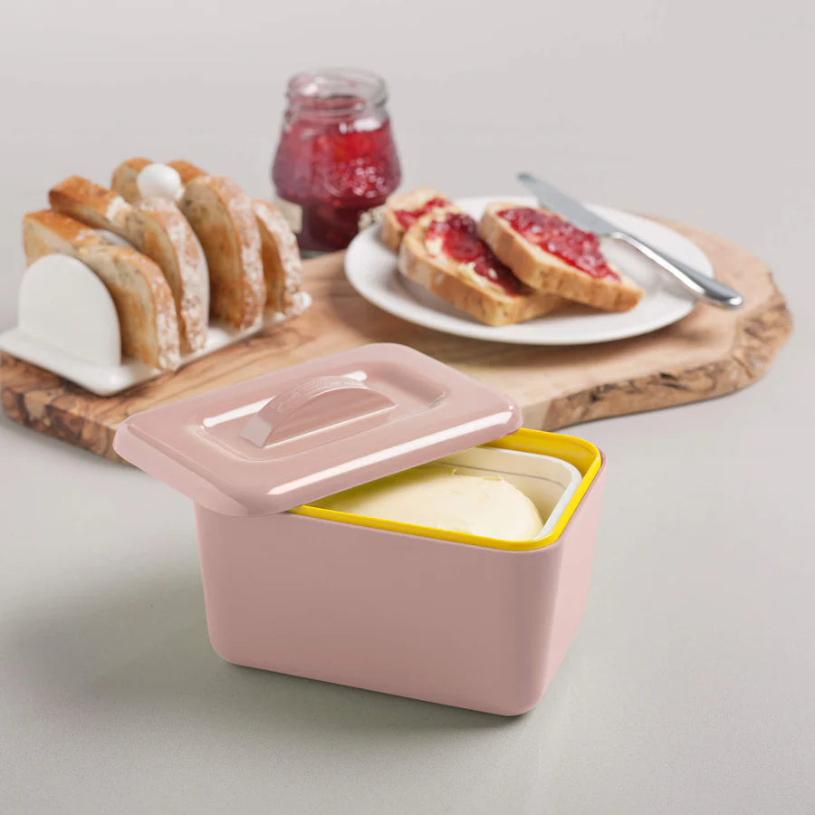 Pink butter dish with a yellow interior on a light gray surface, with toast and jam in the background.